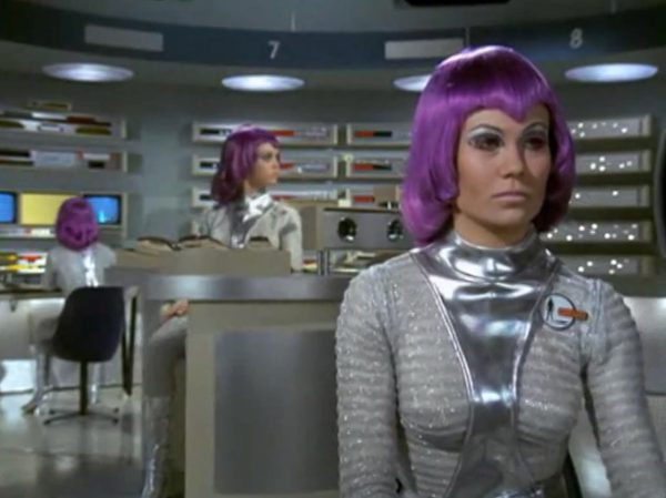 The Moonbase Beauties and Purple Wigs - The women stationed on the SHADO Moonbase are the ultimate icon of retro-chic sci-fi.
The Look - They wore tight, skin-tight metallic silver catsuits or ultra-short metallic miniskirts, complete with matching silver capes, heavy fake eyelashes, and bright, uniform bob-cut purple wigs.
The “Sci-Fi” Excuse - Producer Gerry Anderson claimed that under the harsh fluorescent lights of a lunar base, natural hair would look dull and create static electricity. Therefore, the purple wigs were supposedly “anti-static regulation gear”.
The Reality – The costume designer simply thought purple hair looked incredibly cool against the stark white walls of the moonbase set.
The Contrast - Moonbase Beauties vs. The Boys' Club
The way the show balanced sex appeal with this "advanced space team" is where the corporate logic gets really funny. On one side of the Moonbase, you have the female officers wearing heavy makeup, purple hair, and tight silver miniskirts. They are the ones tracking the alien radar and typing on the computers. But the moment a real fight starts, the show shifts to the three male pilots in their blue pajamas and matching silver motorcycle helmets.
It was a total reflection of 1970 society. The creators wanted the high-fashion, liberal visual hook of the "Moonbase Beauties" to get people to watch the screen, but when it came to the actual space battles, they reverted back to a traditional, serious military setup. The pilots didn't look sexy or futuristic – they looked like a highly trained bobsled team or racecar drivers.
