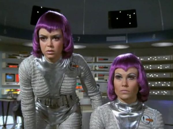 The Moonbase Beauties and Purple Wigs - The women stationed on the SHADO Moonbase are the ultimate icon of retro-chic sci-fi.
The Look - They wore tight, skin-tight metallic silver catsuits or ultra-short metallic miniskirts, complete with matching silver capes, heavy fake eyelashes, and bright, uniform bob-cut purple wigs.
The “Sci-Fi” Excuse - Producer Gerry Anderson claimed that under the harsh fluorescent lights of a lunar base, natural hair would look dull and create static electricity. Therefore, the purple wigs were supposedly “anti-static regulation gear”.
The Reality – The costume designer simply thought purple hair looked incredibly cool against the stark white walls of the moonbase set.
The Contrast - Moonbase Beauties vs. The Boys' Club
The way the show balanced sex appeal with this "advanced space team" is where the corporate logic gets really funny. On one side of the Moonbase, you have the female officers wearing heavy makeup, purple hair, and tight silver miniskirts. They are the ones tracking the alien radar and typing on the computers. But the moment a real fight starts, the show shifts to the three male pilots in their blue pajamas and matching silver motorcycle helmets.
It was a total reflection of 1970 society. The creators wanted the high-fashion, liberal visual hook of the "Moonbase Beauties" to get people to watch the screen, but when it came to the actual space battles, they reverted back to a traditional, serious military setup. The pilots didn't look sexy or futuristic – they looked like a highly trained bobsled team or racecar drivers.