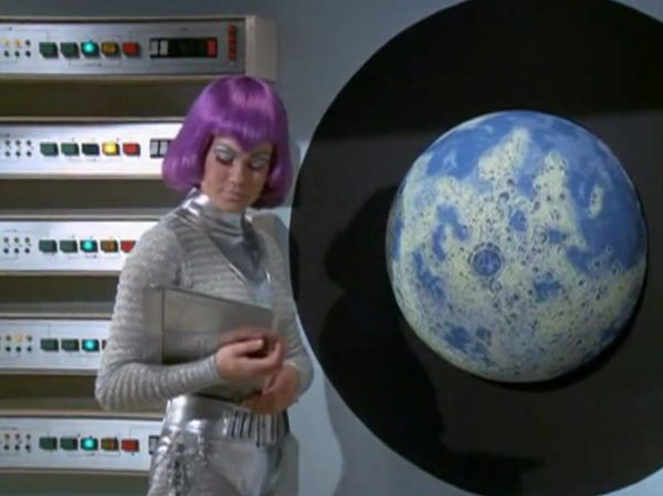 The Moonbase Beauties and Purple Wigs - The women stationed on the SHADO Moonbase are the ultimate icon of retro-chic sci-fi.
The Look - They wore tight, skin-tight metallic silver catsuits or ultra-short metallic miniskirts, complete with matching silver capes, heavy fake eyelashes, and bright, uniform bob-cut purple wigs.
The “Sci-Fi” Excuse - Producer Gerry Anderson claimed that under the harsh fluorescent lights of a lunar base, natural hair would look dull and create static electricity. Therefore, the purple wigs were supposedly “anti-static regulation gear”.
The Reality – The costume designer simply thought purple hair looked incredibly cool against the stark white walls of the moonbase set.
The Contrast - Moonbase Beauties vs. The Boys' Club
The way the show balanced sex appeal with this "advanced space team" is where the corporate logic gets really funny. On one side of the Moonbase, you have the female officers wearing heavy makeup, purple hair, and tight silver miniskirts. They are the ones tracking the alien radar and typing on the computers. But the moment a real fight starts, the show shifts to the three male pilots in their blue pajamas and matching silver motorcycle helmets.
It was a total reflection of 1970 society. The creators wanted the high-fashion, liberal visual hook of the "Moonbase Beauties" to get people to watch the screen, but when it came to the actual space battles, they reverted back to a traditional, serious military setup. The pilots didn't look sexy or futuristic – they looked like a highly trained bobsled team or racecar drivers.