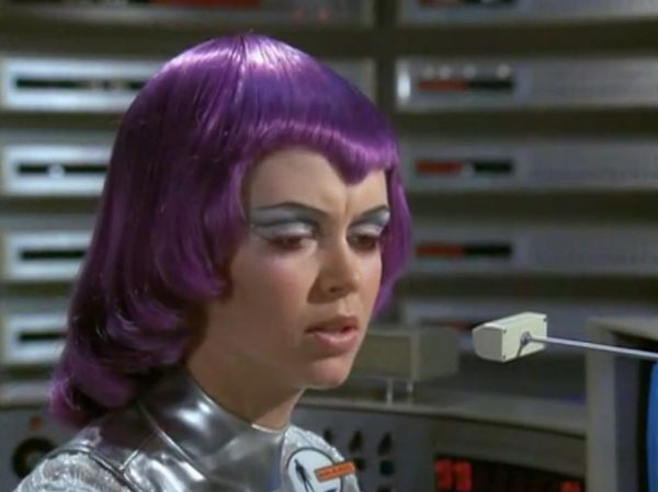 The Moonbase Beauties and Purple Wigs - The women stationed on the SHADO Moonbase are the ultimate icon of retro-chic sci-fi.
The Look - They wore tight, skin-tight metallic silver catsuits or ultra-short metallic miniskirts, complete with matching silver capes, heavy fake eyelashes, and bright, uniform bob-cut purple wigs.
The “Sci-Fi” Excuse - Producer Gerry Anderson claimed that under the harsh fluorescent lights of a lunar base, natural hair would look dull and create static electricity. Therefore, the purple wigs were supposedly “anti-static regulation gear”.
The Reality – The costume designer simply thought purple hair looked incredibly cool against the stark white walls of the moonbase set.
The Contrast - Moonbase Beauties vs. The Boys' Club
The way the show balanced sex appeal with this "advanced space team" is where the corporate logic gets really funny. On one side of the Moonbase, you have the female officers wearing heavy makeup, purple hair, and tight silver miniskirts. They are the ones tracking the alien radar and typing on the computers. But the moment a real fight starts, the show shifts to the three male pilots in their blue pajamas and matching silver motorcycle helmets.
It was a total reflection of 1970 society. The creators wanted the high-fashion, liberal visual hook of the "Moonbase Beauties" to get people to watch the screen, but when it came to the actual space battles, they reverted back to a traditional, serious military setup. The pilots didn't look sexy or futuristic – they looked like a highly trained bobsled team or racecar drivers.